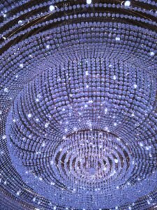 Close-up view of a grand crystal chandelier with rows of sparkling beads and lights reflecting a soft purple glow.